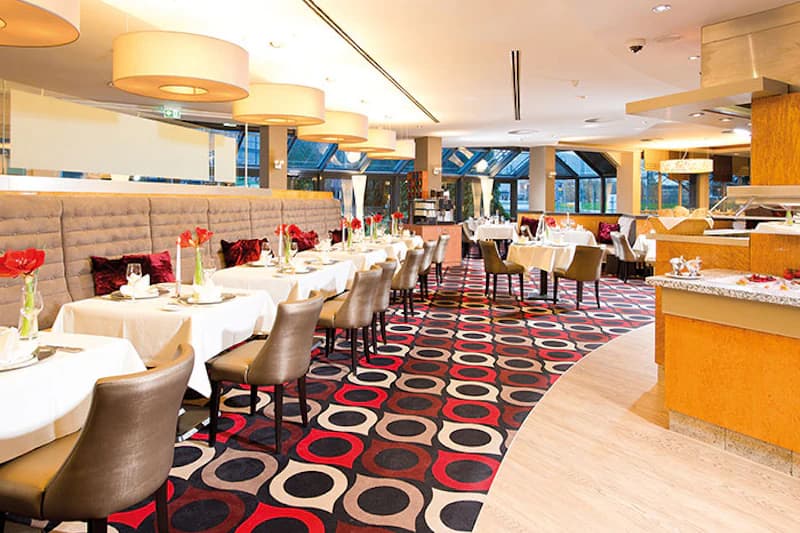 Hoteleigenes Restaurant mit gedeckten Tischen, Stühlen, buntem Kreis-Teppich und Buffetbereich.