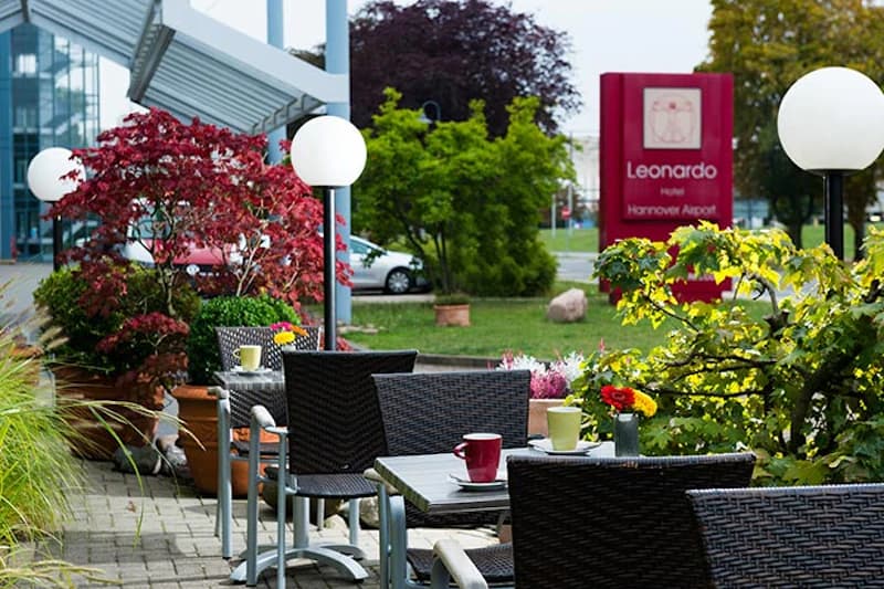 Außenterrasse mit Tischen, Stühlen, Kaffeetassen und Pflanzen vor dem Leonardo Hotel Hannover Airport Schild.