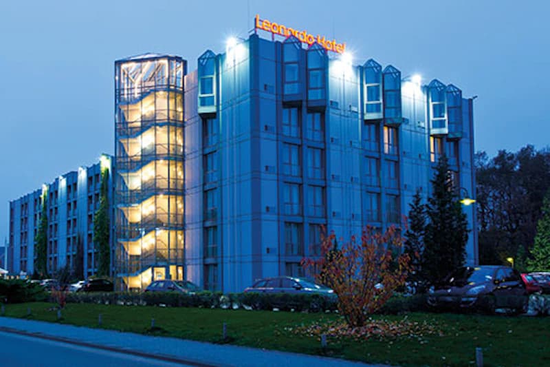 Mehrgeschossiges beleuchtetes Hotelgebäude mit beleuchtetem Außentreppenhaus, Leonardo Hotel Schriftzug und geparkten Autos.