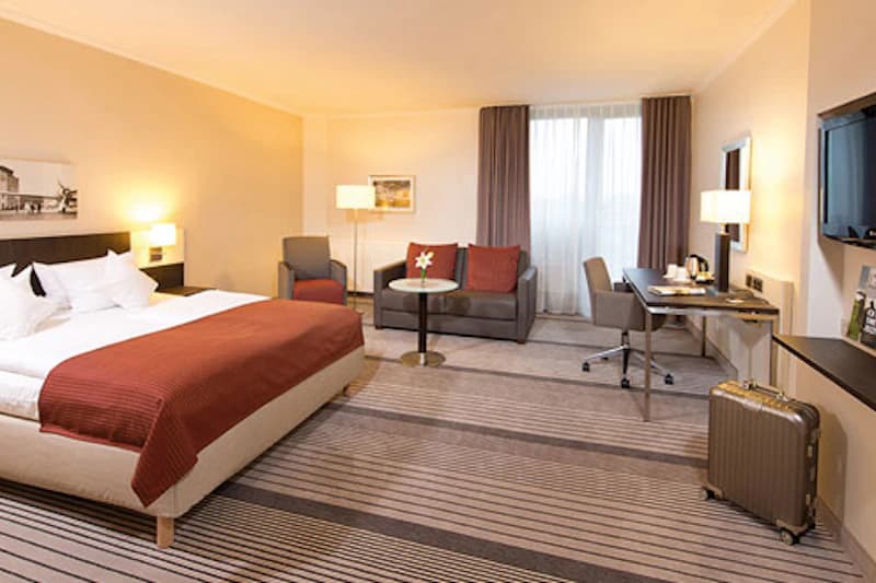 Geräumiges Hotelzimmer mit Doppelbett, Sofa und Sessel, Schreibtisch, Fernseher und einem Koffer.