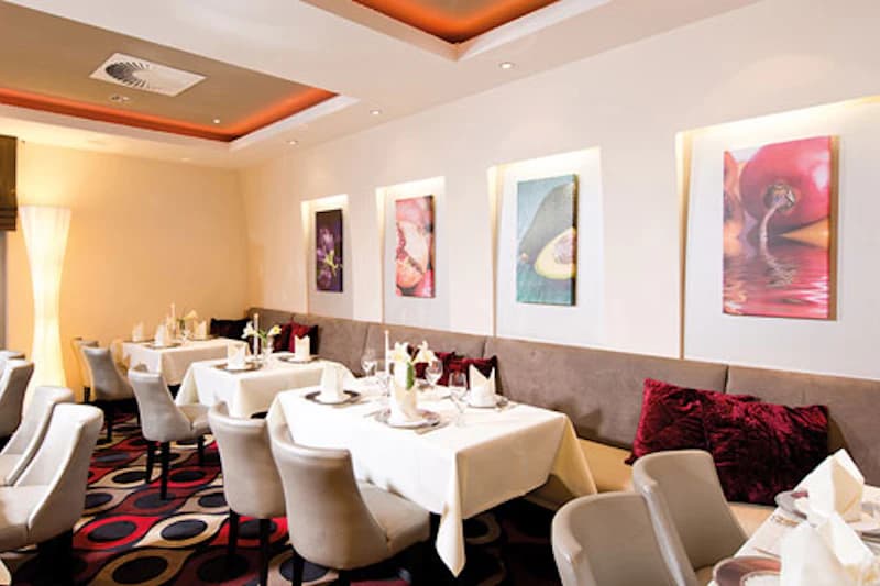 Hotelrestaurant mit gedeckten Tischen, beigen Polsterstühlen, Wandbildern und warmen Deckenleuchten.
