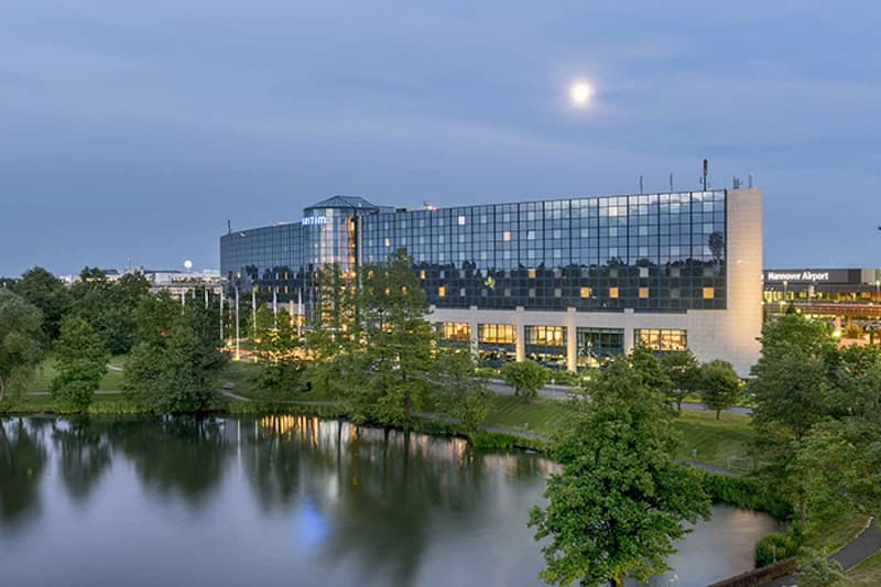 Glasfassade des Maritim Hotels Hannover am Flughafen neben Teich mit Bäumen und Spiegelung bei Dämmerung.