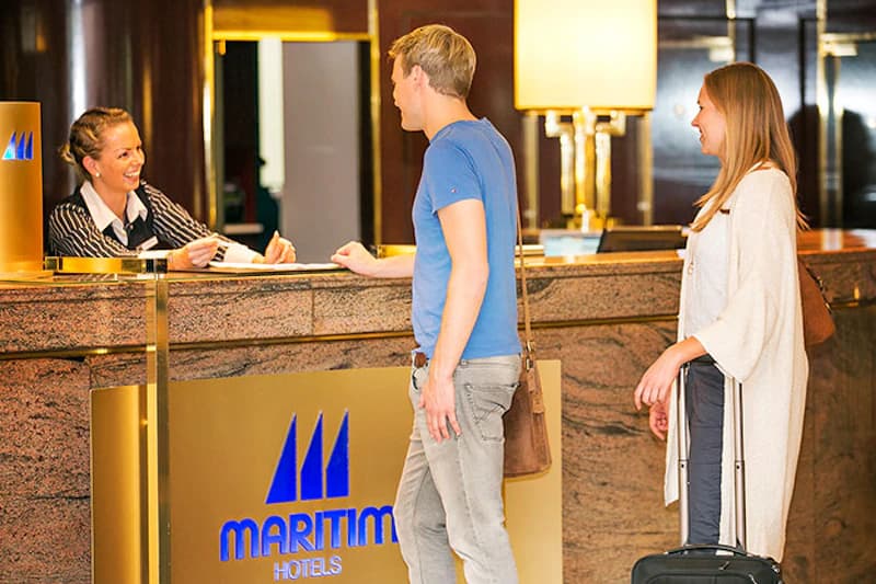 Rezeptionistin reicht Schlüssel an einen Mann am Empfangstresen mit Maritim-Schriftzug, Frau mit Rollkoffer daneben.