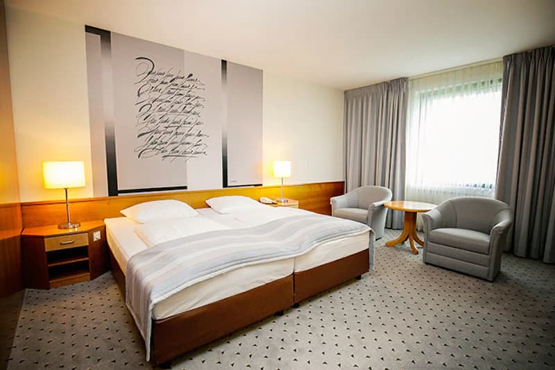 Doppelzimmer mit zwei Betten, Nachttischen und Lampen, zwei Sesseln und rundem Tisch, Fenster mit Vorhängen.