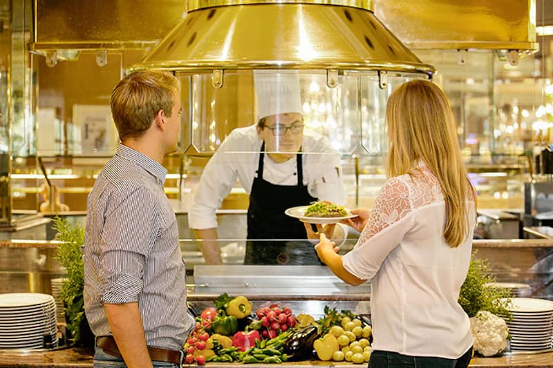Koch überreicht Portion am Buffet an Frau, Mann wartet, Theke mit frischem Gemüse.