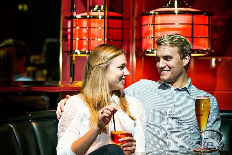 Paar sitzt in roter Hotelbar auf Lederbank, Frau hält Cocktail mit Strohhalm, Mann hält Bierglas.