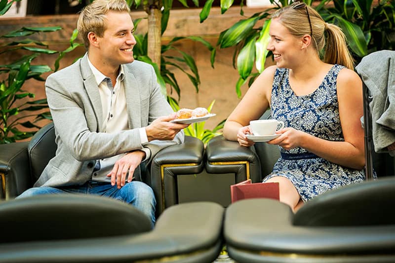 Zwei lächelnde Gäste in Lounge, Mann reicht Teller mit Gebäck, Frau hält Tasse, Pflanzen im Hintergrund.