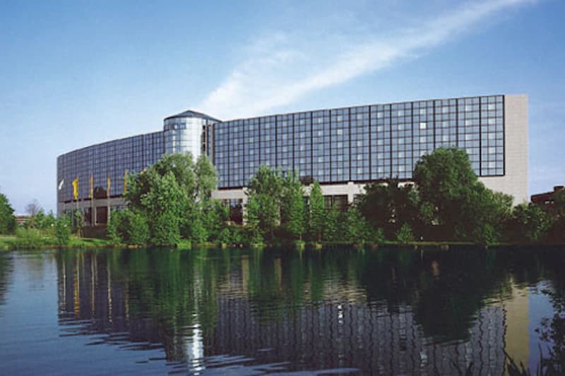 Großes geschwungenes Hotelgebäude mit Glaspaneelen, runder Glasvorbau, Bäume am Ufer und Spiegelung im See.