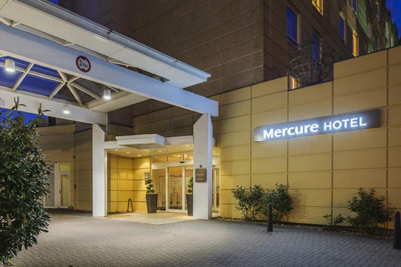 Eingang des Mercure Hotels mit überdachter Zufahrt, Leuchtschild, Glastüren und Pflanzen bei Dämmerung.