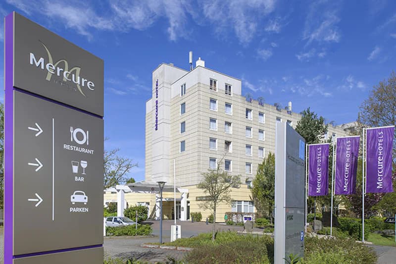 Hotelgebäude mit Eingangsüberdachung, großem Hinweisschild für Restaurant, Bar und Parken und lila Mercure-Fahnen.