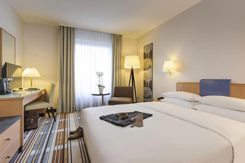 Helles Hotelzimmer mit Doppelbett, Schreibtisch, Fernseher, Sessel und Fenster mit Vorhängen; Kleidung und Schuhe auf Bett und Boden.