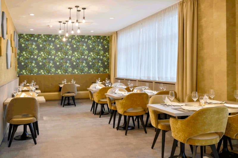Heller Hotelrestaurant mit gedeckten Tischen, gelben Samtstühlen, großem Fenster und floraler Tapete.