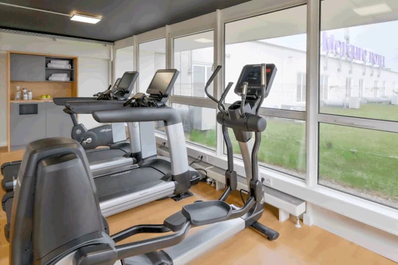 Fitnessraum mit zwei Laufbändern und Crosstrainer vor großen Fenstern, Mercure-Schriftzug außen sichtbar.
