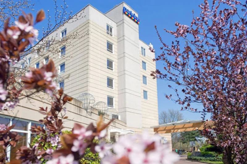 Sechsstöckiges Hotelgebäude mit farbigem Logo, blühende Sträucher im Vordergrund und blauem Himmel.