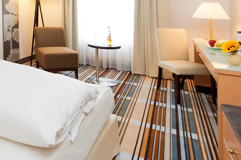 Hotellzimmer mit Bettvorderkante, gestreiftem Teppich, Sessel, kleinem runden Tisch mit Flasche und Schreibtisch mit Stuhl.