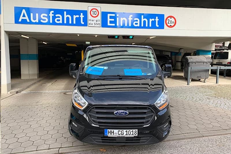 Schwarzer Ford-Van vor Parkhauseinfahrt mit blauen Zetteln auf dem Armaturenbrett und Schildern Ausfahrt und Einfahrt.