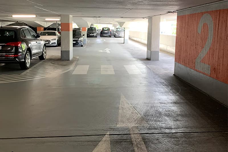 Parkhausinnenraum mit Fahrbahnmarkierungen, Bodenpfeilen, Zebrastreifen, geparkten Autos und großer Zahl 2 an der Wand.