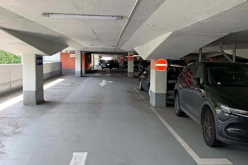 Innenansicht eines Parkdecks mit geparkten Autos rechts, Pfeilmarkierungen auf dem Boden und Säulen mit Einfahrtsverbotsschildern.