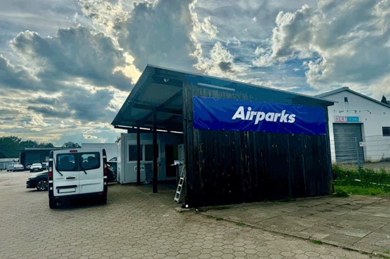 Holzunterstand mit blauem Airparks-Banner neben geparktem weißem Van auf gepflastertem Parkplatz, wolkiger Himmel.
