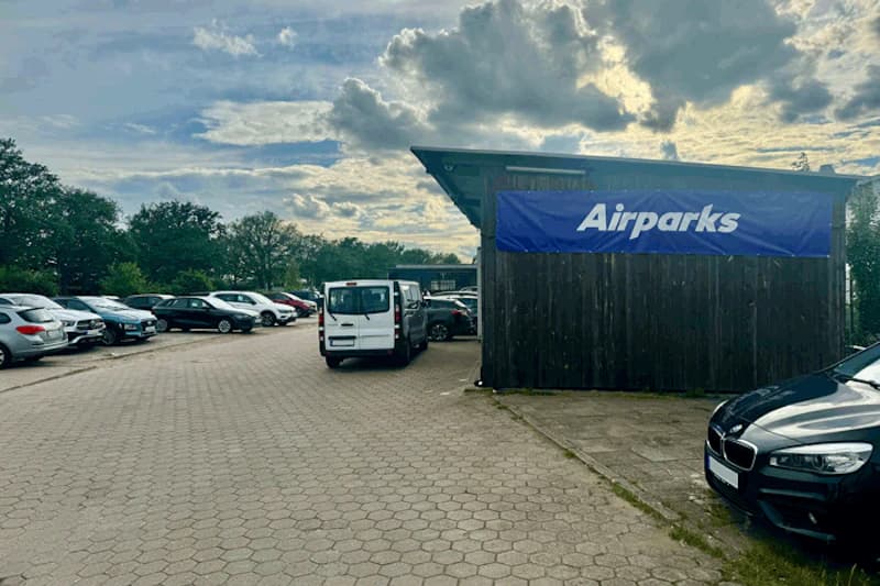 Gepflasterter Parkplatz mit geparkten Autos und Holzgebäude mit blauem Airparks-Banner unter bewölktem Himmel.