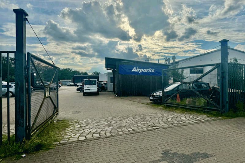 Offenes Metalltor zu einem Parkplatz mit geparkten Autos und Airparks-Banner am Gebäude unter bewölktem Himmel.