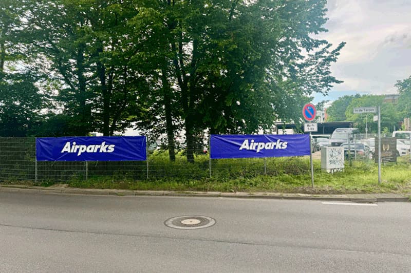Zwei blaue Airparks-Banner an einem Zaun vor Bäumen und geparkten Fahrzeugen, Straße im Vordergrund.