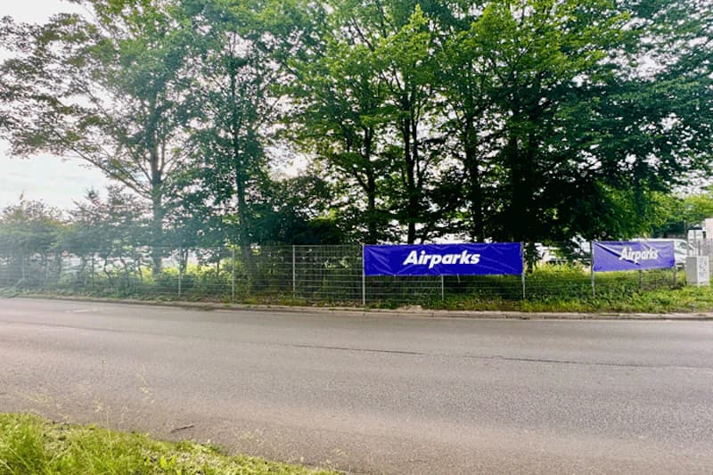 Leere Straße vor Metallzaun mit blauen Airparks-Bannern und Bäumen im Hintergrund.