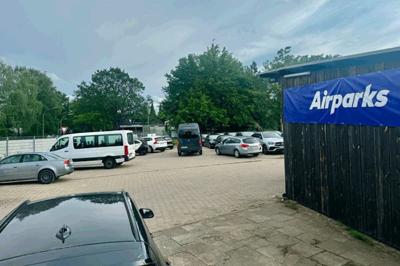 Parkplatz mit geparkten Autos und Kleinbus, Holzzaun mit Airparks-Banner, gepflasterte Fläche und Bäume im Hintergrund.