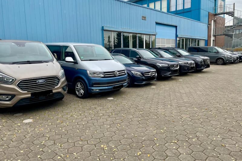 Reihe geparkter Autos vor blauem Industriegebäude auf gepflastertem Parkplatz.