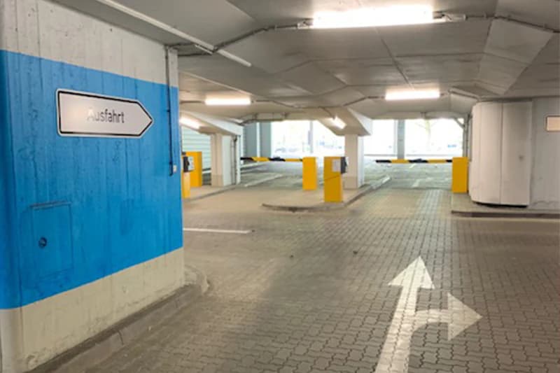 Parkhausinnenraum mit blauer Säule und Ausfahrt-Schild, Bodenpfeil und gelben Schrankenautomaten.