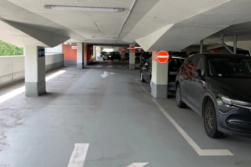 Überdachtes Parkdeck mit Fahrspur, Bodenpfeilen, geparkten Autos rechts und Durchfahrtsverbotsschildern an Betonpfeilern.