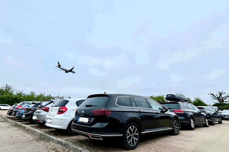 Reihe geparkter Autos auf offenem Freiparkplatz, im Himmel ein anfliegendes Flugzeug mit Lufthansa-Heck.