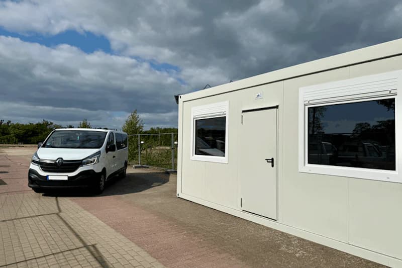 Weißer Transporter neben weißem Containerbüro auf gepflastertem Platz vor Zaun und wolkenverhangenem Himmel.