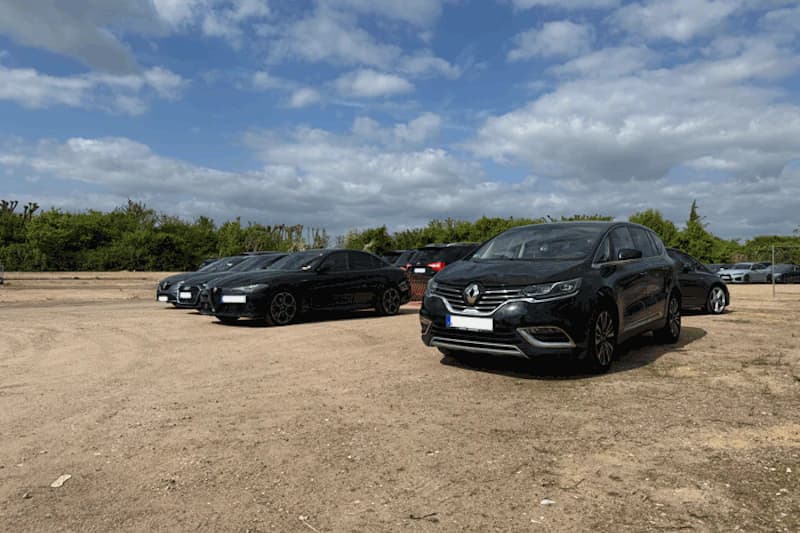 Unbefestigter Parkplatz mit mehreren geparkten Autos, im Vordergrund schwarzer Renault, Bäume und bewölkter Himmel.