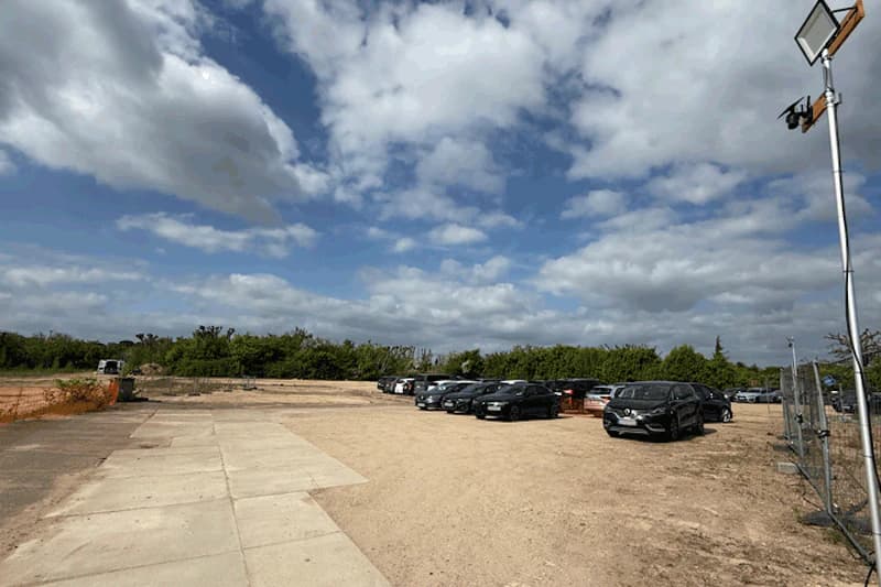 Offener Schotterparkplatz mit geparkten Autos rechts, Sicherheitszaun und Kameramast rechts, Baumreihe im Hintergrund.