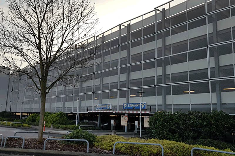 Mehrstöckiges Parkhaus mit Metallfassade, Einfahrtsschildern, kahlem Baum und randnaher Bepflanzung.