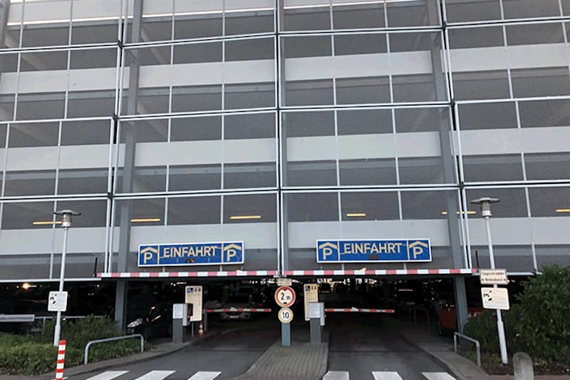 Mehrstöckiges Parkhaus mit Metallfassade, zwei Einfahrten, Schranken, Höhenbegrenzung und Zebrastreifen.