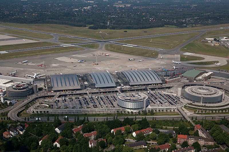 Luftaufnahme des Flughafens Hamburg mit Terminalgebäuden, Rollfeld, mehreren Flugzeugen und runden Parkhäusern im Vordergrund.