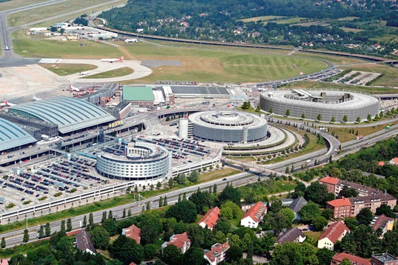 Luftaufnahme des Flughafens Hamburg mit Terminalhallen, drei runden Parkhäusern, Rollfeld und angrenzenden Straßen.