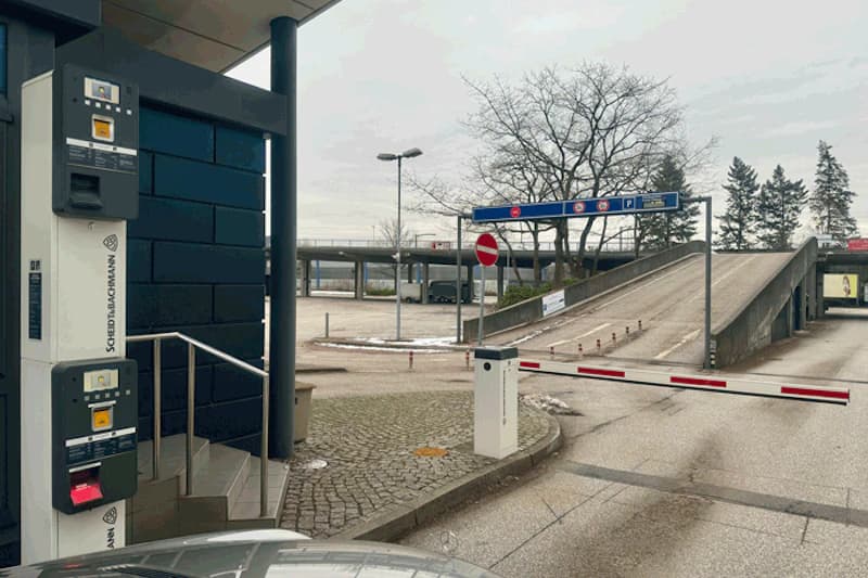 Schranke und Parkscheinautomaten am Zufahrtbereich eines Parkdecks mit Rampe und Verkehrsschildern.