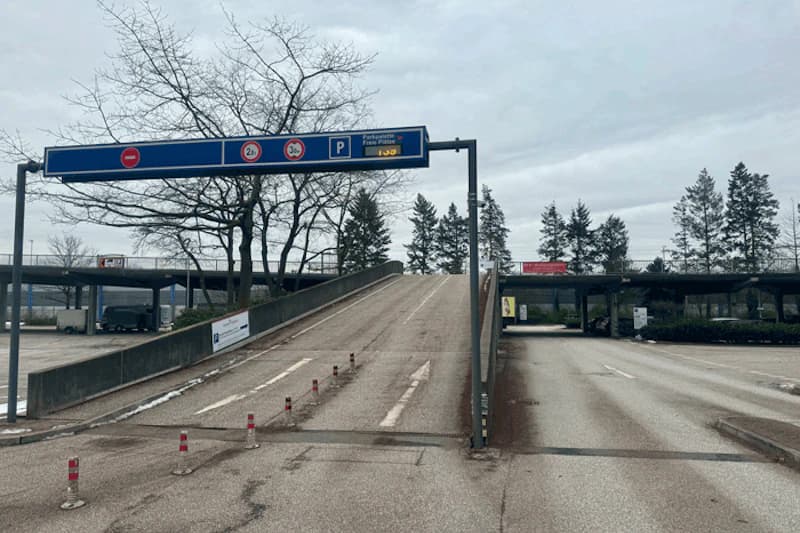 Zufahrt zu einer Betonrampe des Parkdecks mit blauem Verkehrszeichen, Absperrpfosten und kahlen Bäumen.