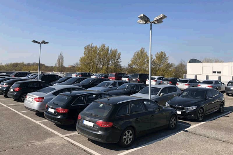Parkdeck mit mehreren geparkten Pkw, Beleuchtungsmasten und Bäumen im Hintergrund.