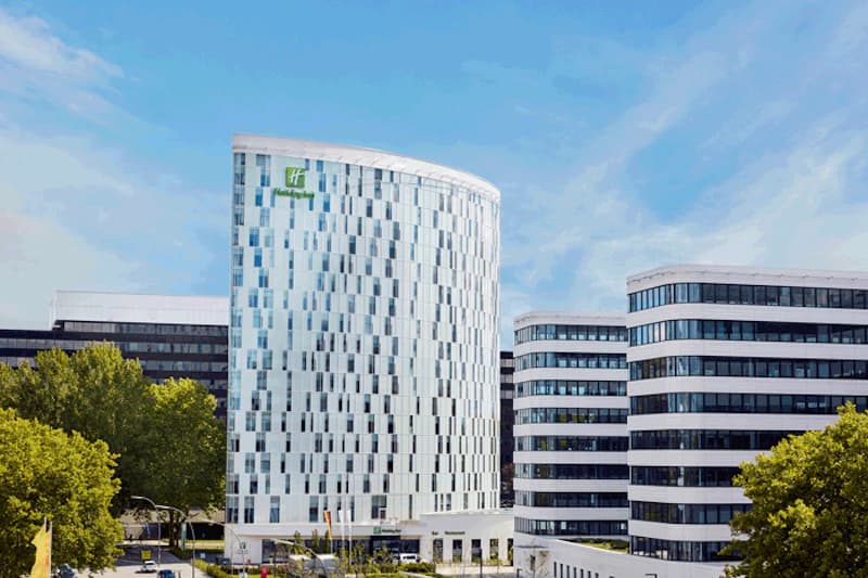 Hohes gebogenes Hotelgebäude mit Holiday Inn Logo, daneben mehrgeschossige Bürogebäude und Bäume unter blauem Himmel.