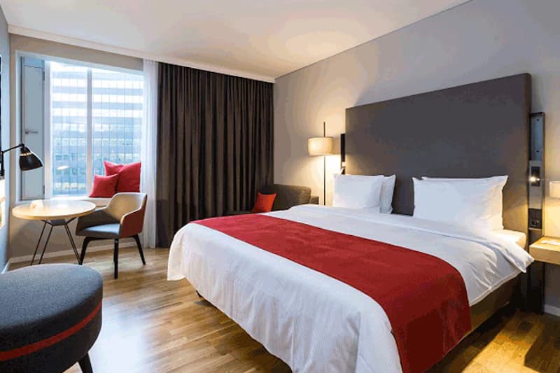 Hotelzimmer mit großem Doppelbett, rotem Betttuch, Sitzbereich am Fenster und Holzfußboden.