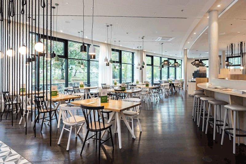 Heller Hotelrestaurant mit Holztafeln, gemischten Stühlen, Hängeleuchten und Bartheke vor großen Fenstern.