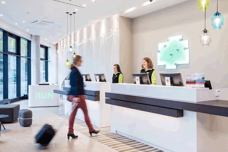 Hotelrezeption mit Tresen, zwei Empfangsmitarbeiterinnen und vorbeigehender Gast mit Rollkoffer.