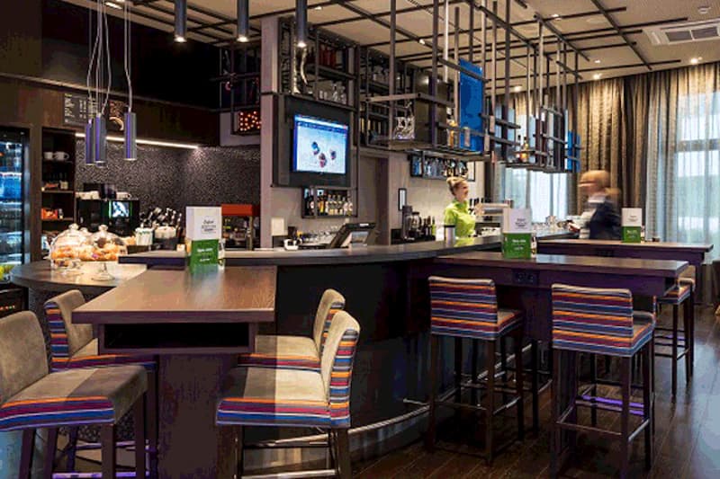 Hotelbar mit Tresen, gestreiften Barhockern, hohen Tischen und zwei Mitarbeitenden im Hintergrund.