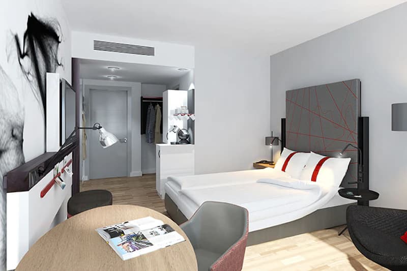 Modernes Hotelzimmer mit Doppelbett, gemustertem Kopfteil, Beistelltisch, Esstisch mit Stühlen und Garderobe.