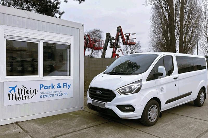 Weißer Moin Moin Transporter neben weißem Container mit Park & Fly Schild, Kräne im Hintergrund.