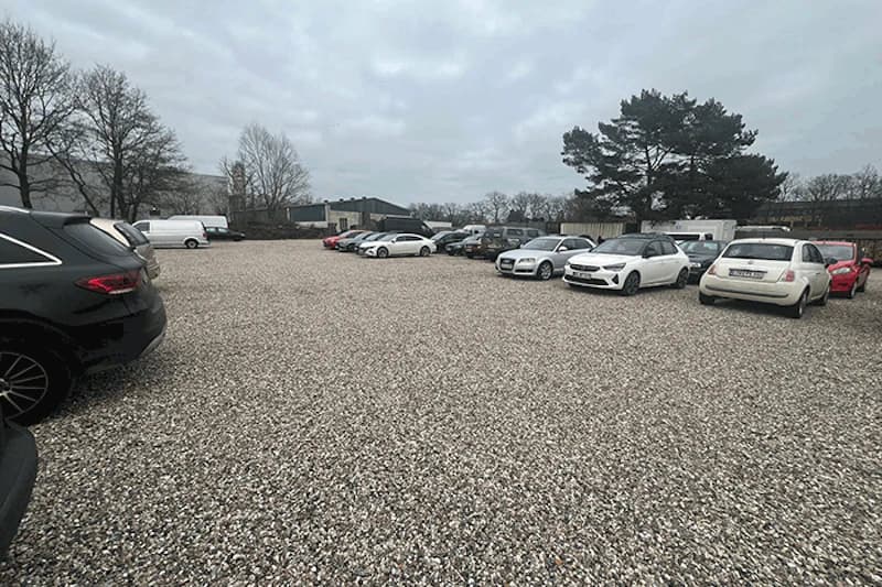 Schotterparkplatz mit mehreren geparkten Autos, Bäumen im Hintergrund und wolkigem Himmel.